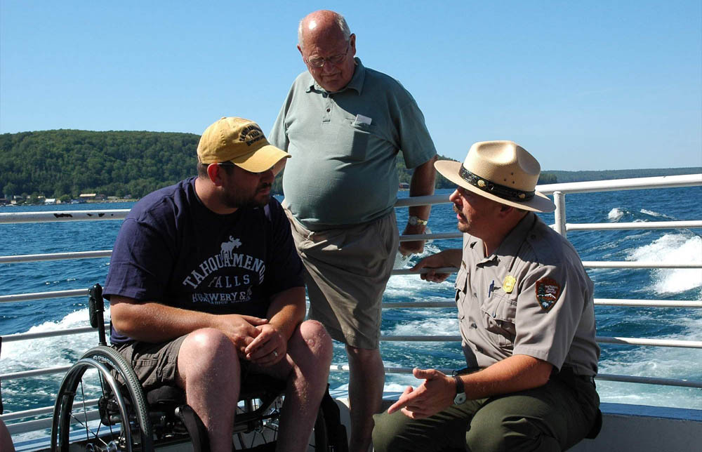 Man in NPS uniform with a shield-shaped badge and broad rim hat kneels on the deck of a boat talking to a man in wheelchair and a standing man.